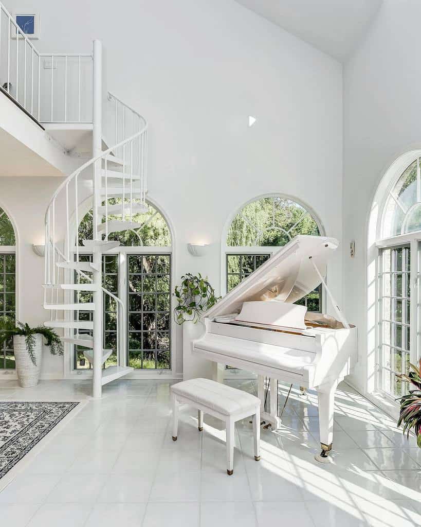 A spacious white room with a grand piano, spiral staircase, large arched windows, and indoor plants