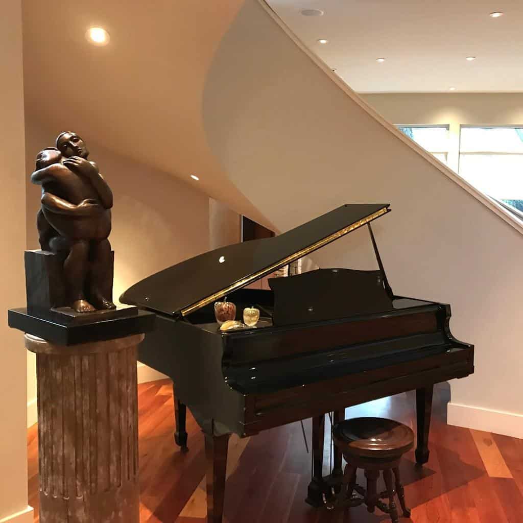 A grand piano with a closed lid and stool sits beside a modern sculpture on a column in a warmly lit room with wooden floors