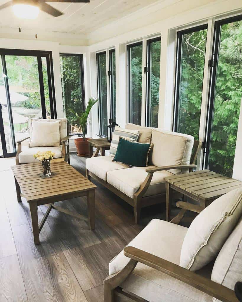 Cozy sunroom with wooden furniture, beige cushions, a small coffee table with flowers, and tall windows overlooking greenery