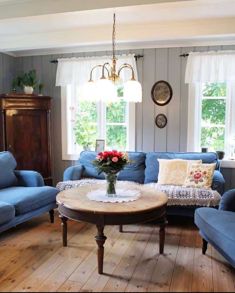 Round wooden coffee table with turned legs, styled with a lace doily and a vase of fresh flowers, set in a cozy cottage-style living room with blue sofas