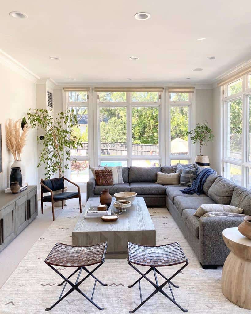 Bright living room with large windows, gray sectional sofa, wooden coffee table, woven chairs, and indoor plants