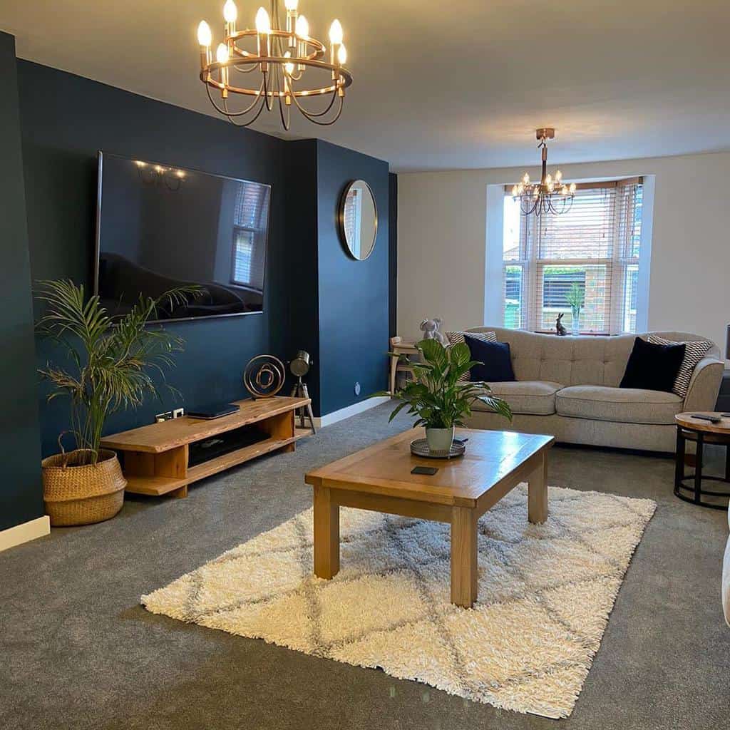 Modern living room with a large TV, gray sofa, wooden coffee table on a white rug, plants, and a chandelier; dark accent wall with a mirror