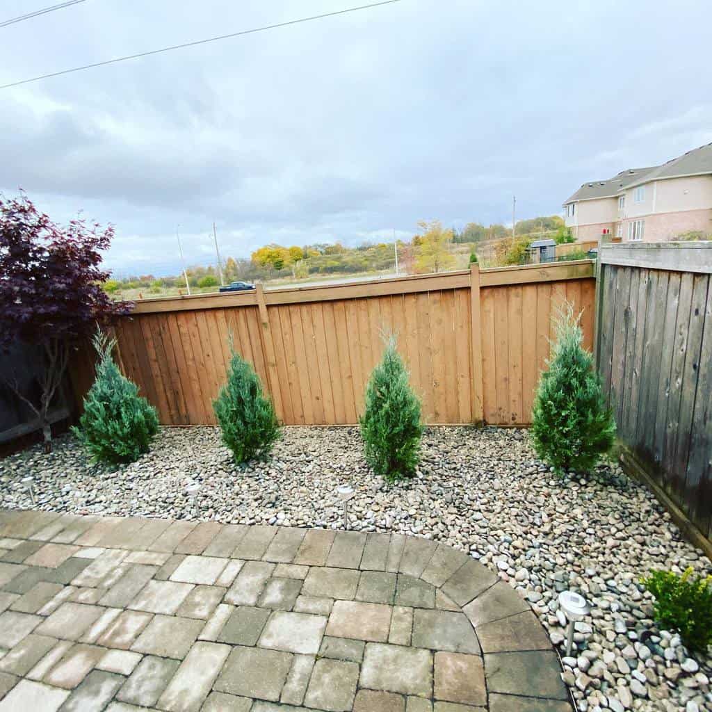 Backyard xeriscape with evergreen juniper shrubs, a wooden fence, decorative rocks, and a paved patio under a cloudy sky