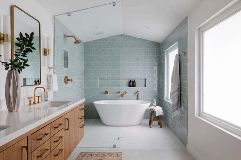 A modern wet room with a freestanding tub, light blue tiles, double vanity, brass fixtures, and a large window