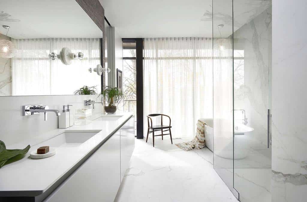 Minimalist wet room with white marble, large mirror, glass shower, and freestanding tub; a chair and sheer curtains complete the look