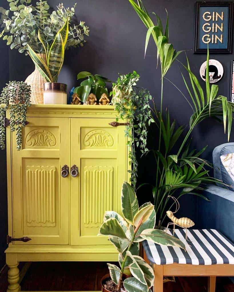 Bold yellow vintage cabinet against a dark wall, surrounded by lush green plants, decorative accents, and a striped side table