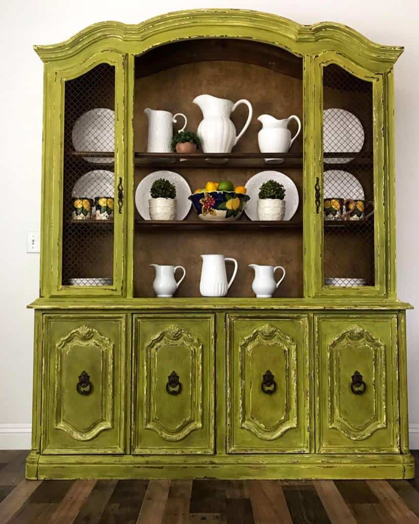 Vintage hutch painted distressed olive green, showcasing white pitchers, decorative plates, and lemon-themed accents on wooden shelves