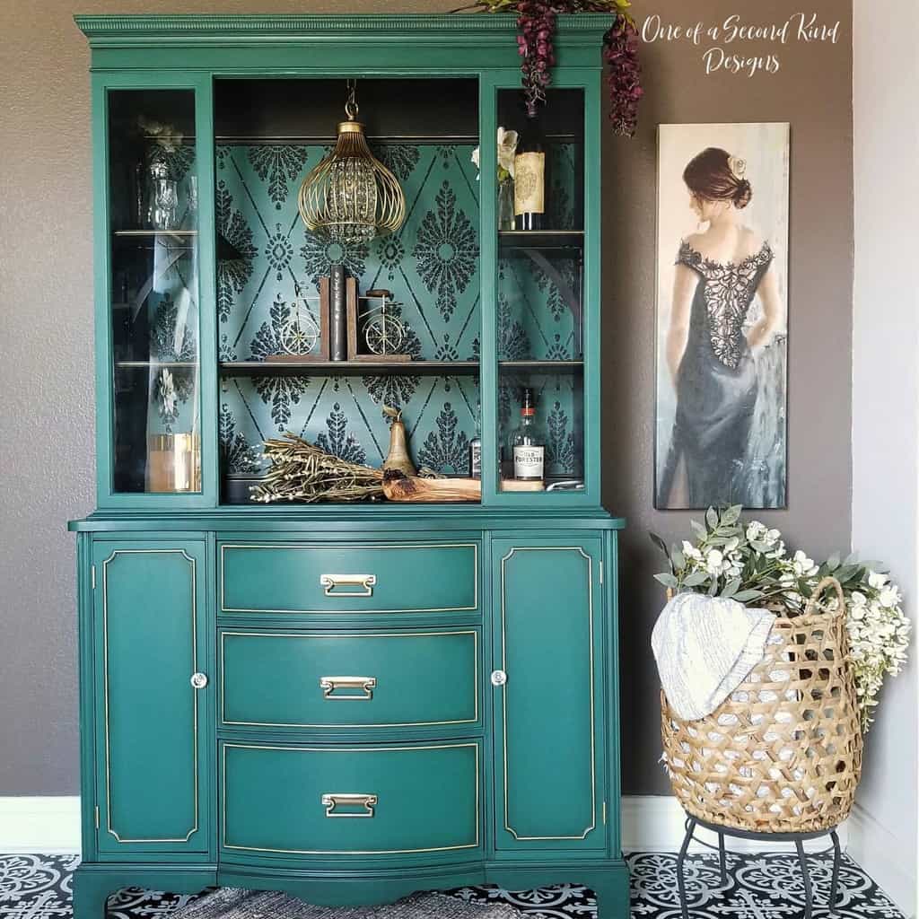 Elegant teal hutch with gold accents, glass doors, and a patterned backdrop, styled with books, décor, and a vintage chandelier