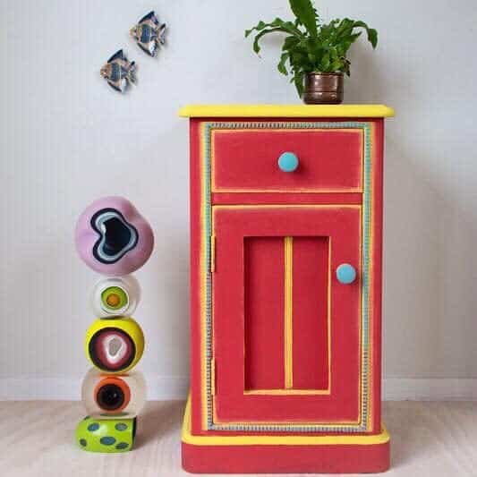 Bold red and yellow chalk-painted cabinet with blue knobs, paired with eclectic stacked decor and a potted plant on top