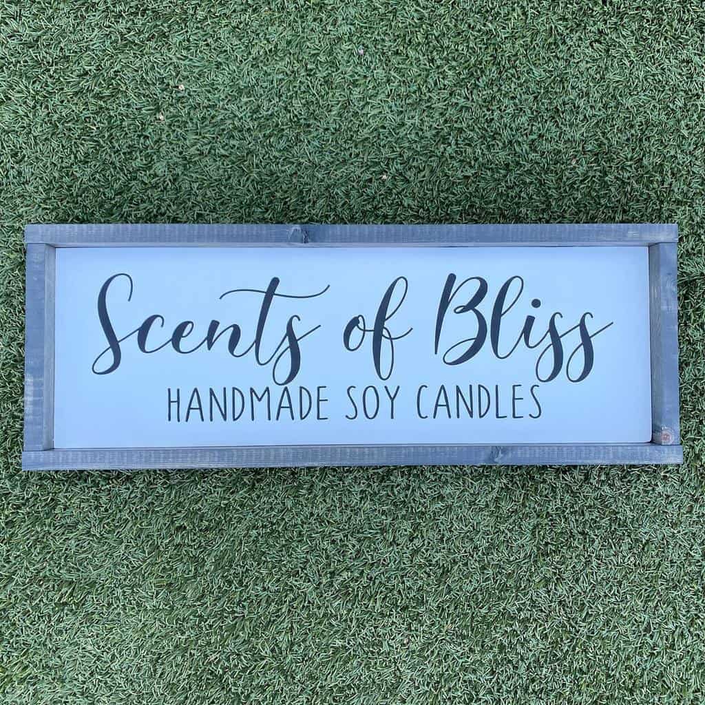 A wooden sign on grass reads "Scents of Bliss Handmade Soy Candles