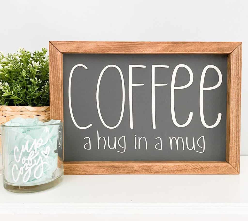 Framed wooden sign reading "Coffee: a hug in a mug" next to a green plant and a glass jar with mint packets