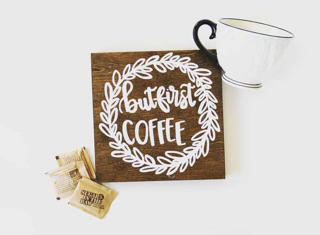 A wooden sign with "but first, coffee" in white next to a white coffee cup and sugar packets on a white background