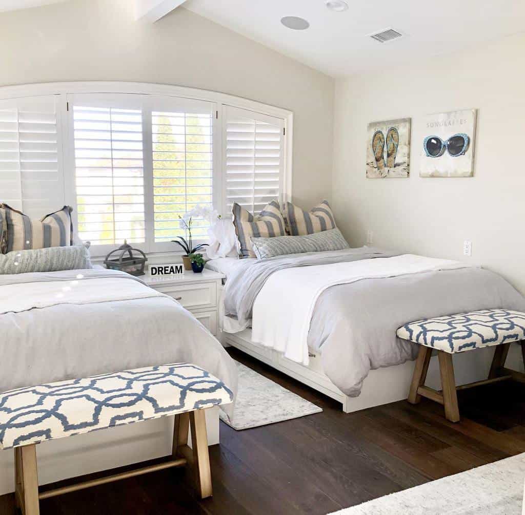 Bright bedroom with two beds, striped pillows, decorative benches, and art on walls. White shutters let in natural light; cozy vibe