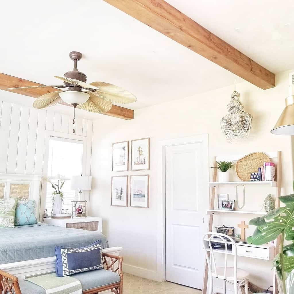 Bright bedroom with wooden beams, ceiling fan, bed, desk, and wall art; decor includes plants and wicker accents