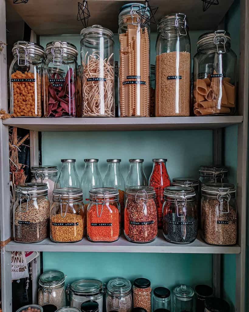 A pantry shelf with various dried foods like pasta, legumes, and grains stored in glass jars with black labels