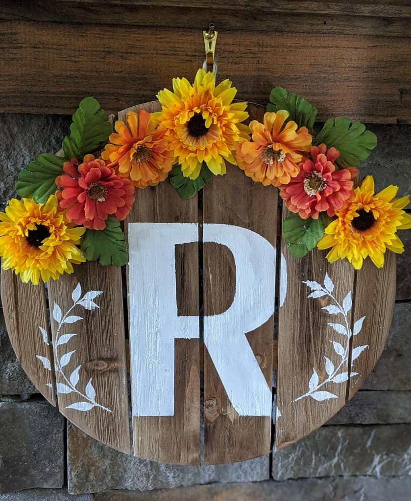 Wooden sign with a large white letter "R," adorned with yellow and orange flowers and leaves at the top, hanging on a stone wall