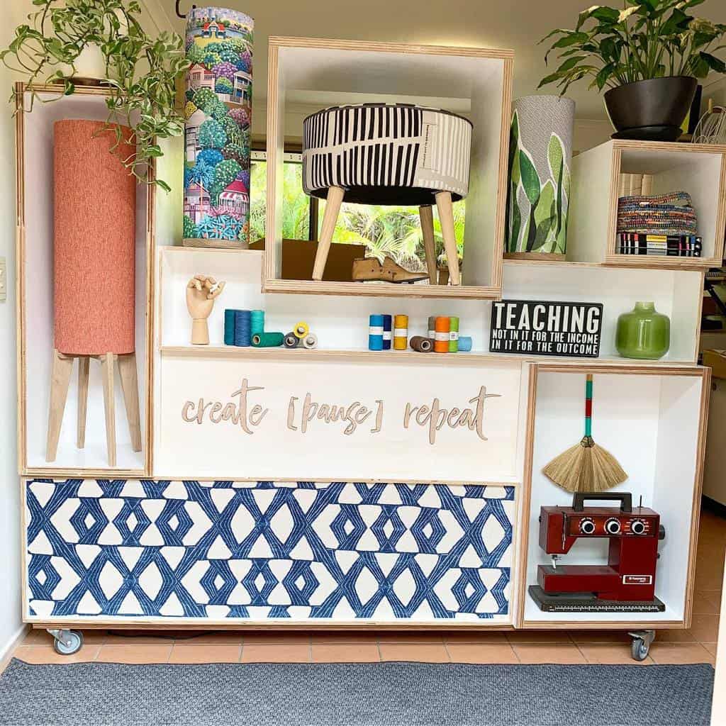 Colorful, eclectic bookshelf with plants, fabric rolls, a striped stool, craft supplies, and a red typewriter: "Create [Pause] Repeat" text