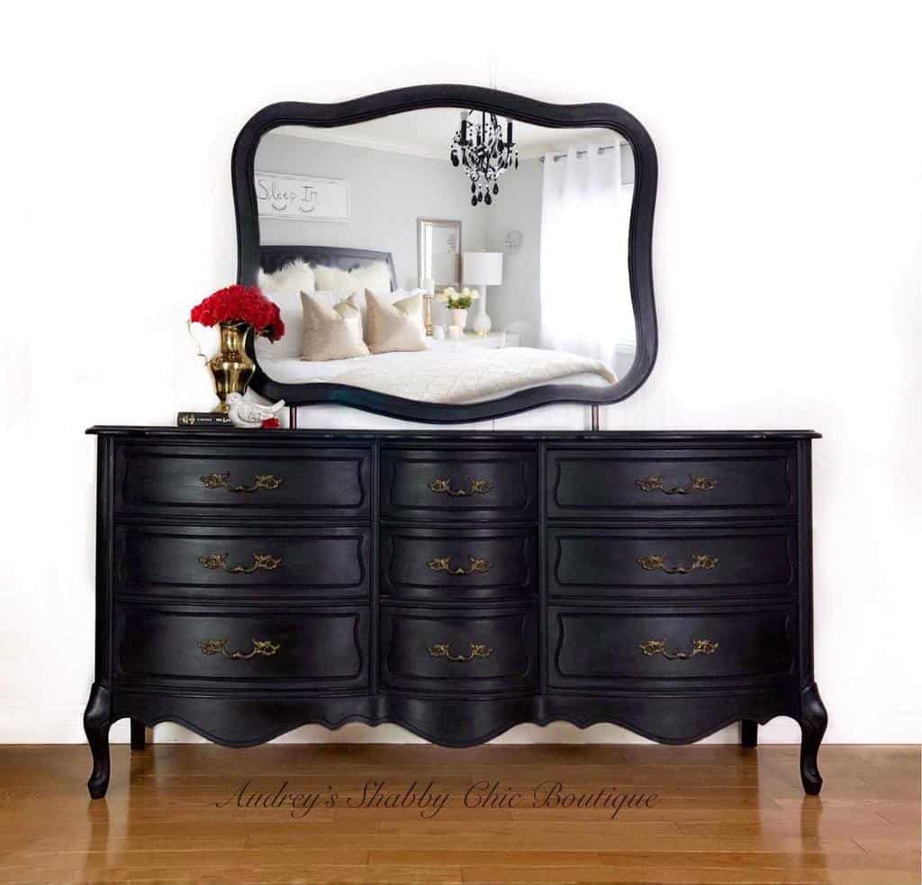 Black chalk-painted vintage dresser with ornate gold handles, a curved mirror, and red floral decor, reflecting a cozy bedroom setting