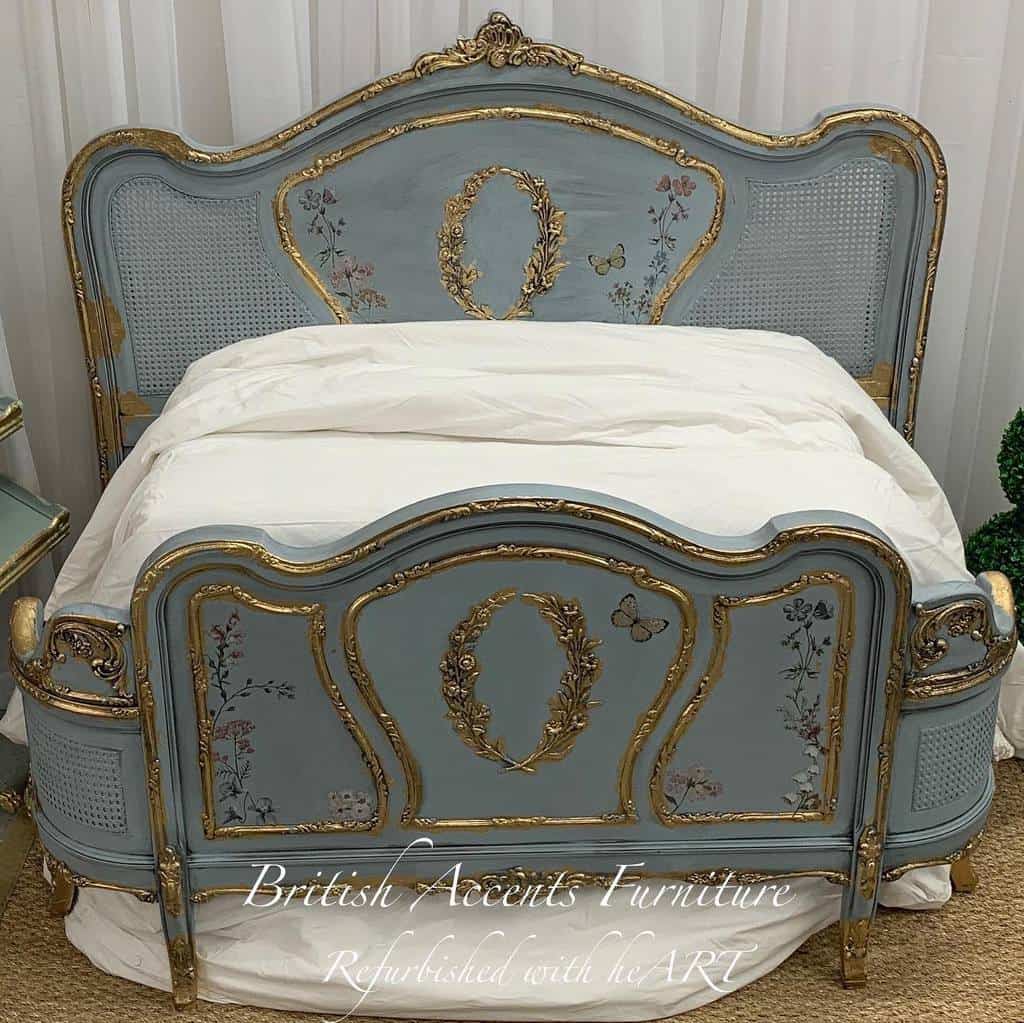 Elegant vintage bed frame painted in muted blue with gold detailing, floral accents, and cane panels, styled with white bedding