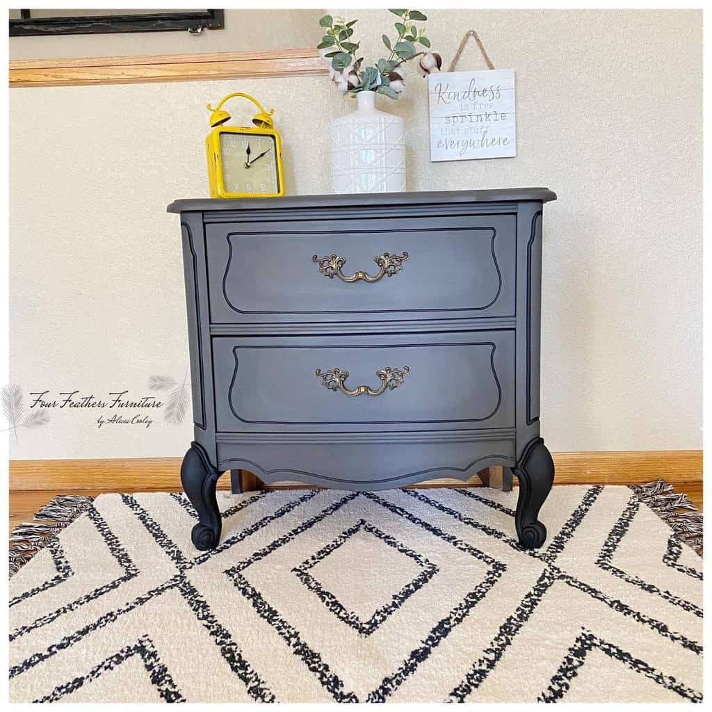 Chic dark gray chalk-painted vintage nightstand with ornate gold handles, styled with a yellow clock, vase, and decor on a patterned rug