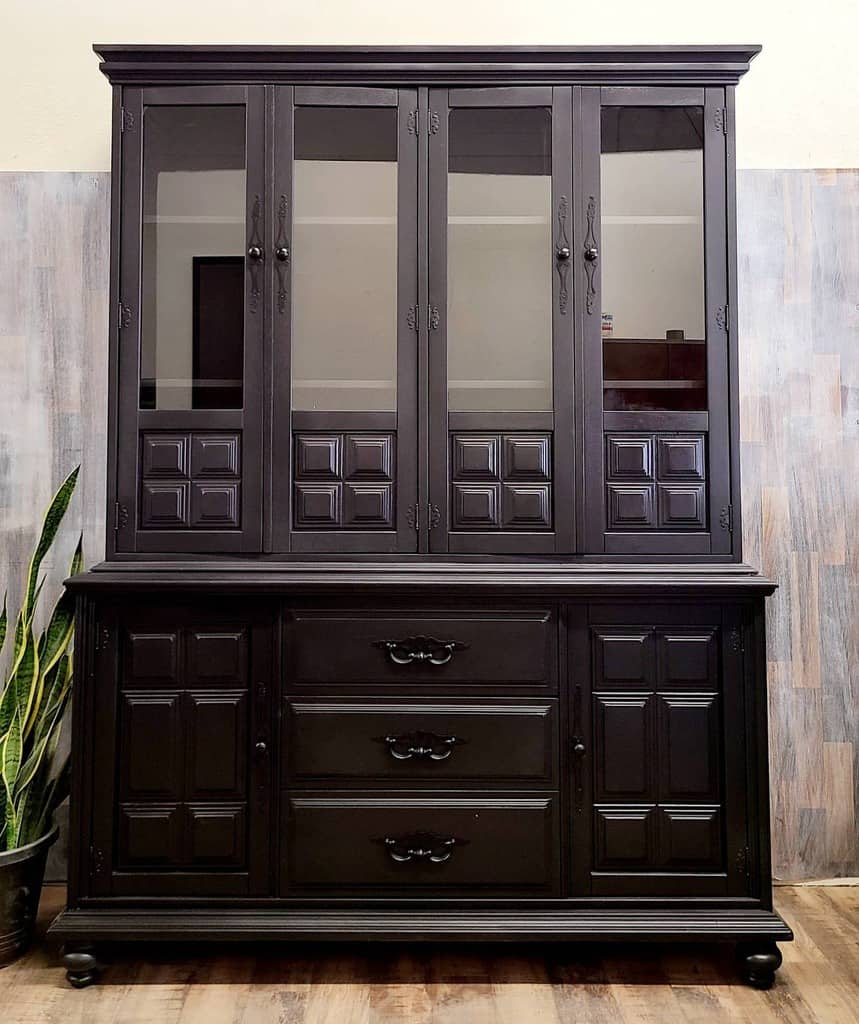 Large black chalk-painted hutch with glass-paneled doors, intricate wood detailing, and vintage-style handles, set against a rustic backdrop