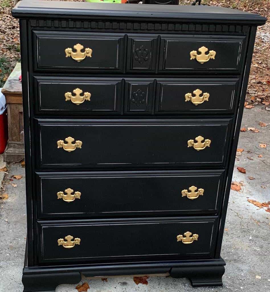 Black chalk-painted vintage dresser with gold handles and intricate wood detailing, set outdoors on a rustic surface with fallen leaves