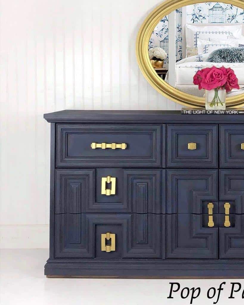 Navy blue chalk-painted dresser with gold geometric handles, styled with a round gold mirror, pink roses, and a white-paneled backdrop