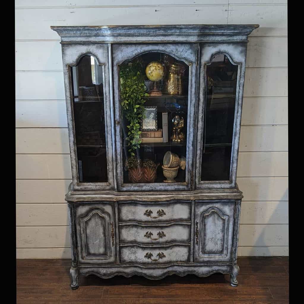 Vintage china cabinet with a distressed gray chalk paint finish, gold hardware, glass doors, and antique decor inside