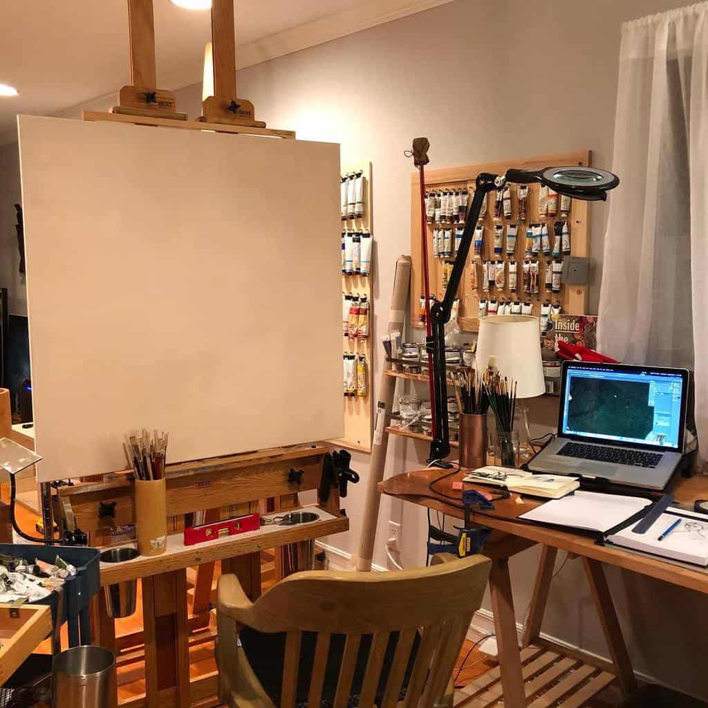 Artist's workspace with a wooden easel, blank canvas, paint tubes, brushes, laptop, and desk in a well-lit room