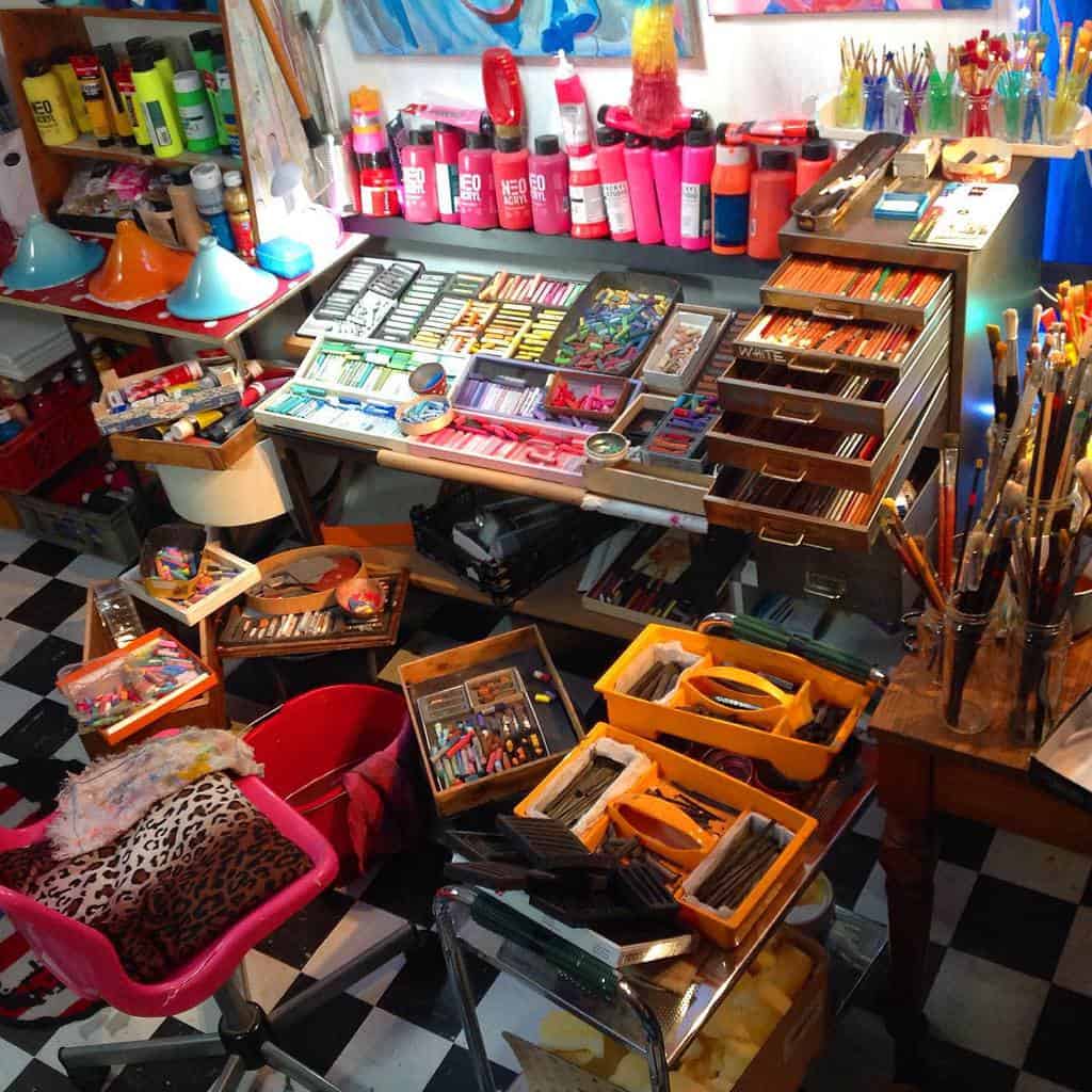 Colorful art studio with paints, brushes, and various art supplies on a checkered floor under bright lighting that highlights the vibrant materials