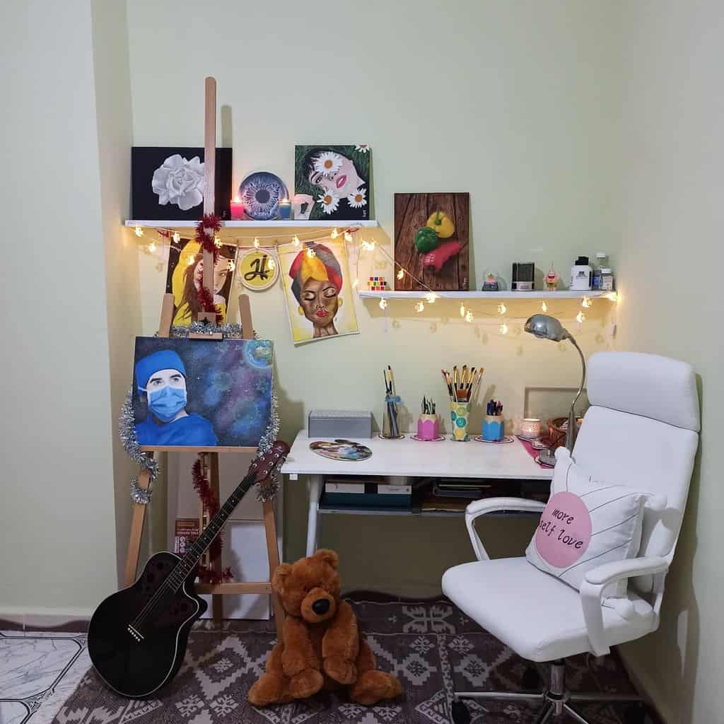 Cozy art workspace with paintings, fairy lights, guitar, teddy bear, and a white chair with a "more self love" pillow