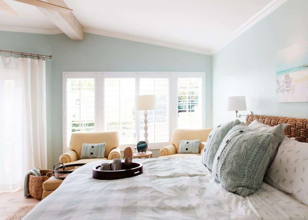 A bright bedroom with a large bed, knit pillows, two armchairs, and a window with sheer curtains allowing natural light to fill the room