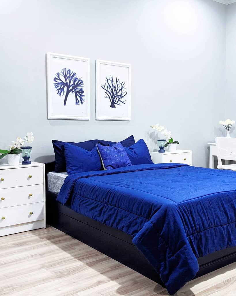 Bedroom with a dark blue bedspread, white walls, and two tree-themed artworks, as well as white bedside tables and a desk