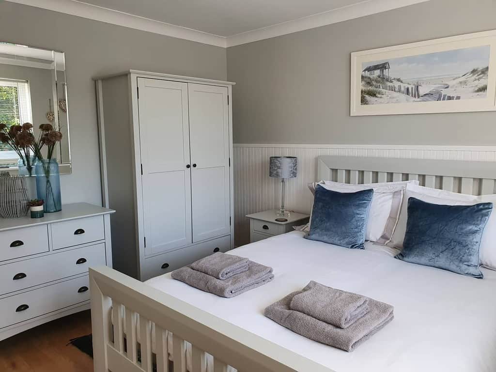 Coastal bedroom with a double bed, gray and blue decor, a dresser with vases, a wardrobe, two folded towels, and a landscape picture