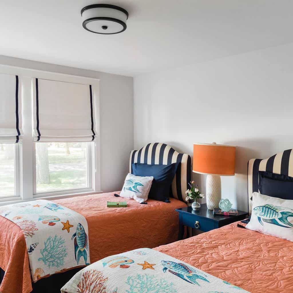 Bright bedroom with two twin beds, striped headboards, and ocean-themed quilts, featuring an orange lamp and a window with light filtering shades