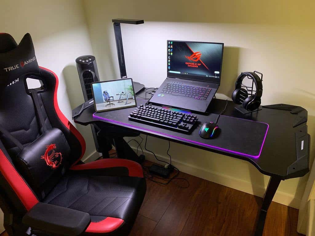 Gaming setup with a laptop on a black desk, a mechanical keyboard, mouse, tablet, headphones, and a black and red gaming chair