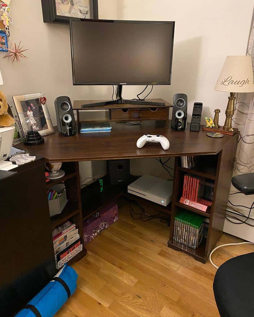 Gaming setup with a monitor, speakers, and a white controller on a wooden desk, surrounded by books and a lamp