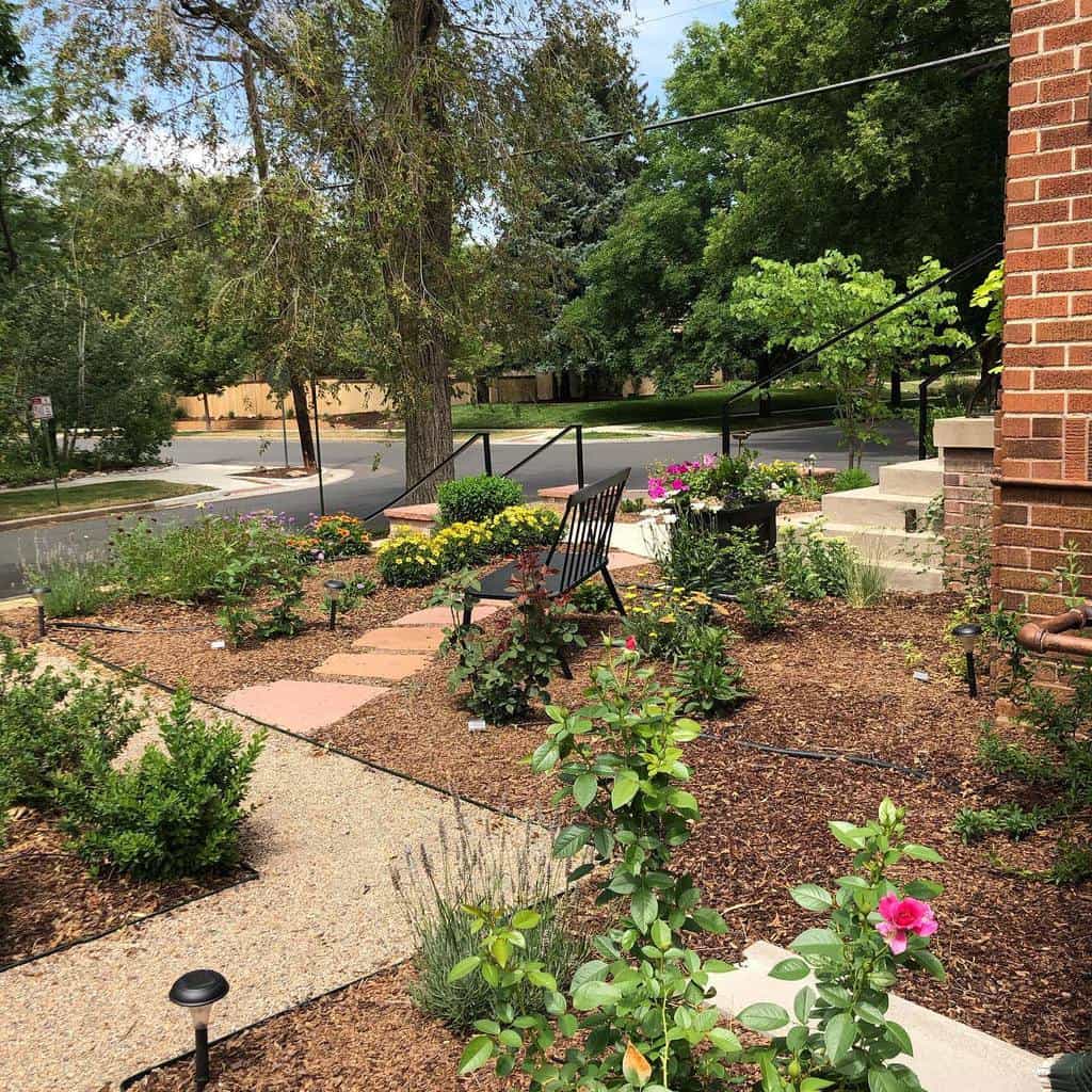 Charming front yard xeriscape with flowering plants, mulch pathways, and a seating area, surrounded by lush greenery and a brick home