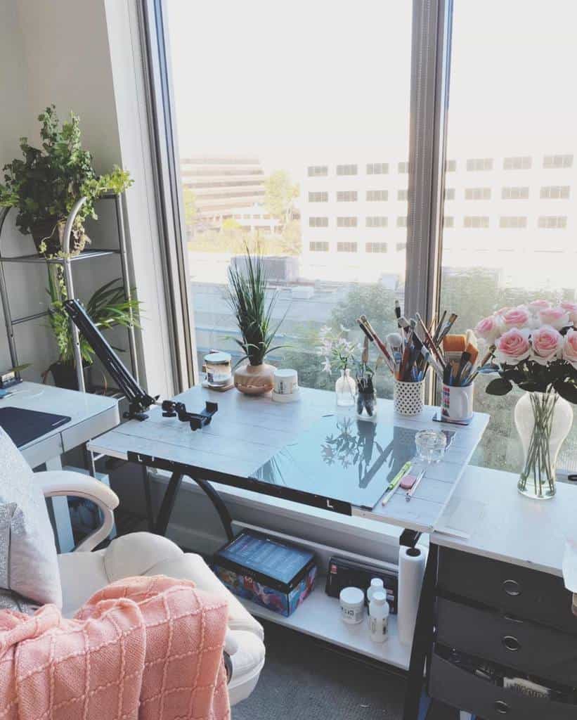 Bright art studio with large window, desk filled with brushes, supplies, and vase of pink roses; cozy chair with pink blanket