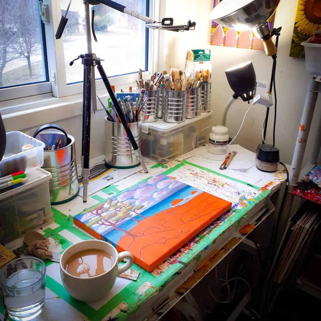 Art studio workspace with a painting in progress, brushes, tripod, and a cup of coffee near a window