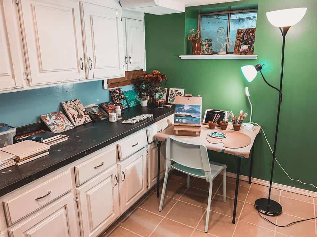 Cozy art studio with white cabinets, green walls, easel on a desk, chair, and floor lamp; art supplies and paintings around the room