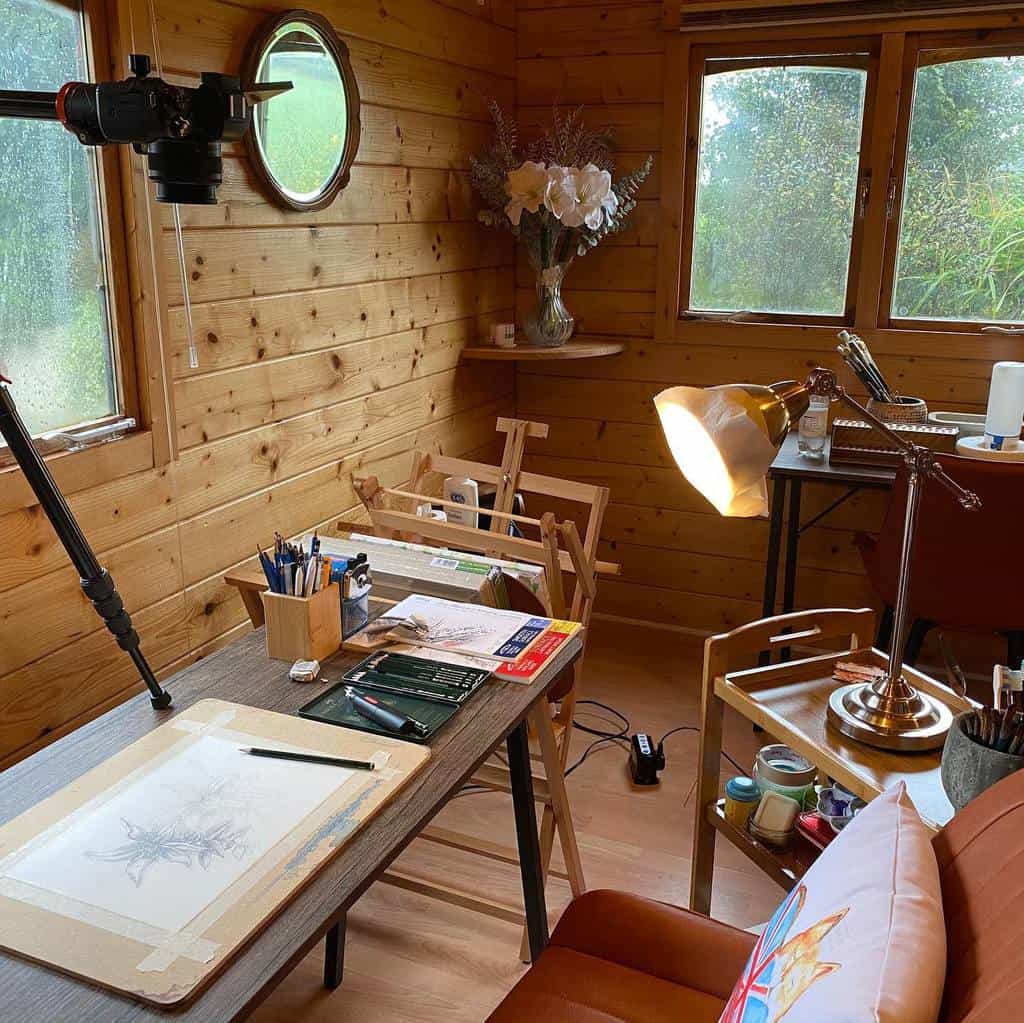 Cozy art studio with wooden walls, desk with drawing tools, lamp, and a tripod. Large windows show greenery outside