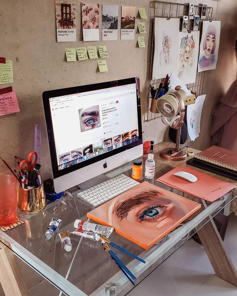 Artistic workspace with computer, painting of an eye, art supplies, and inspiration board with sticky notes and sketches