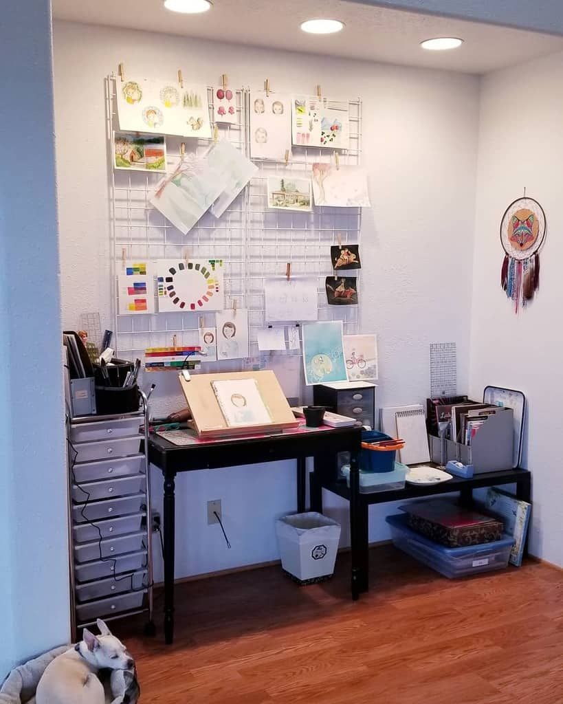 Artist's workspace with drawings on a wire grid, a desk with supplies, storage bins, and a colorful dreamcatcher on the wall