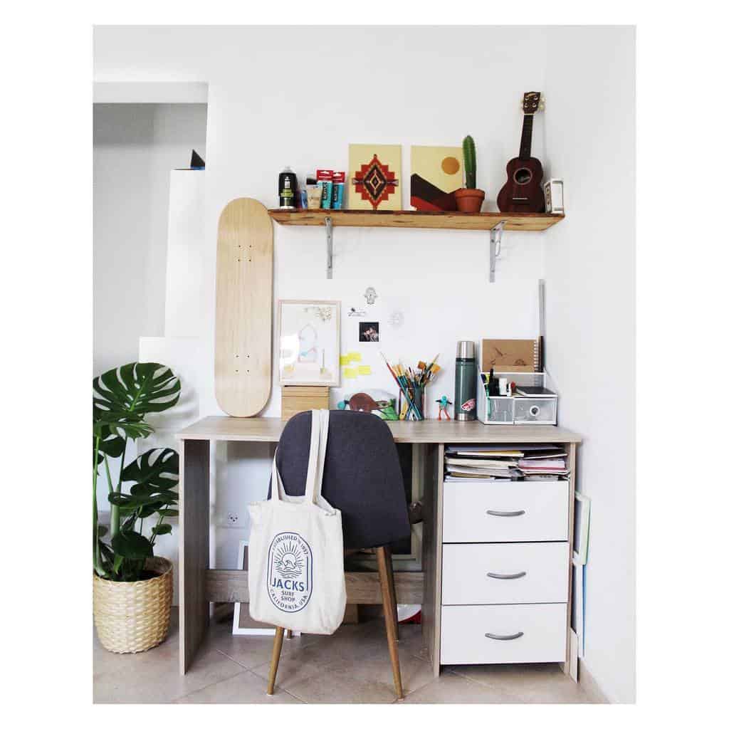 Minimalist workspace with a wooden desk, chair, skateboard, and shelf with books and plant; tote bag hangs on the chair's back