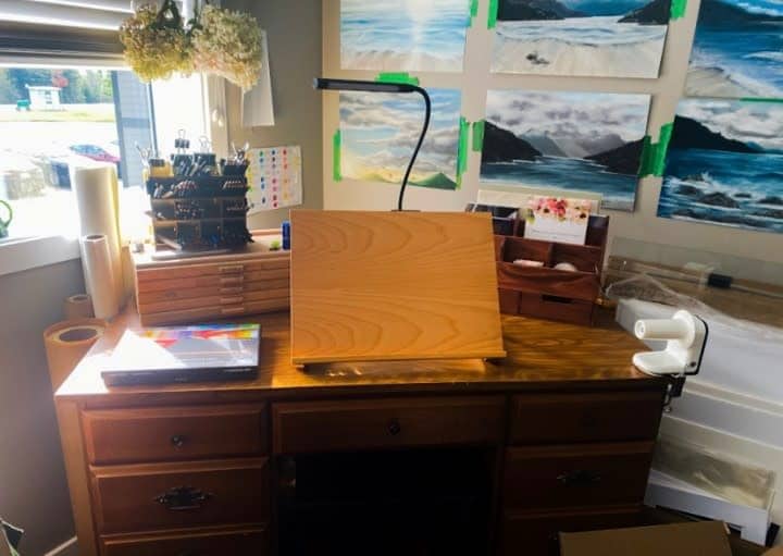 Art studio corner with an easel, paint supplies, and landscape paintings on the wall, brightly lit by a desk lamp