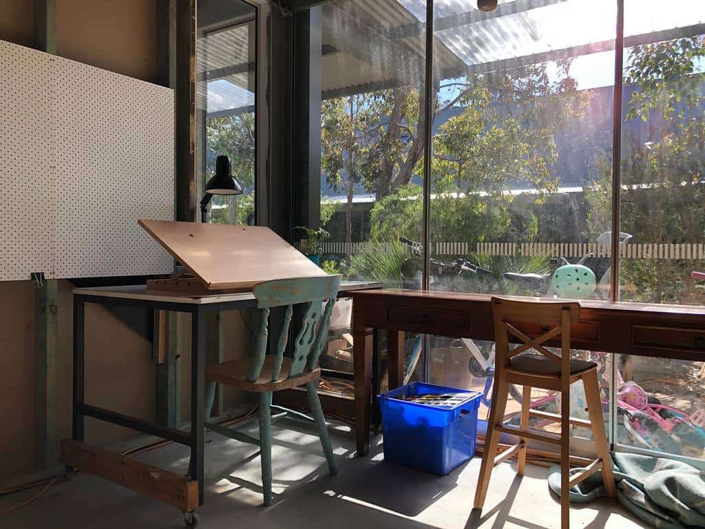 Sunny art studio with a drafting table, two chairs, a blue box, and large windows overlooking a garden with trees