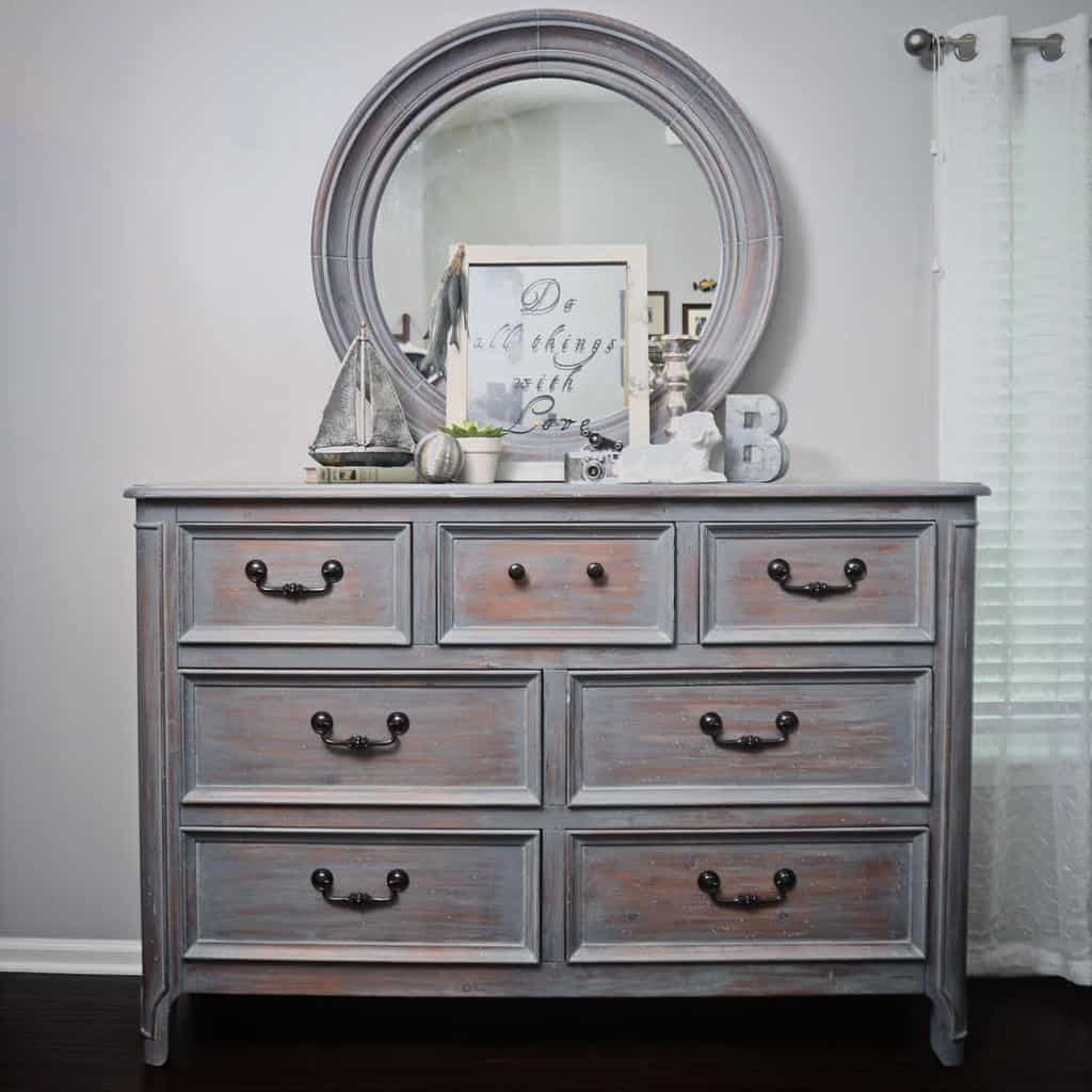 Rustic gray chalk-painted dresser with a distressed finish, black metal handles, and a round mirror, styled with nautical decor