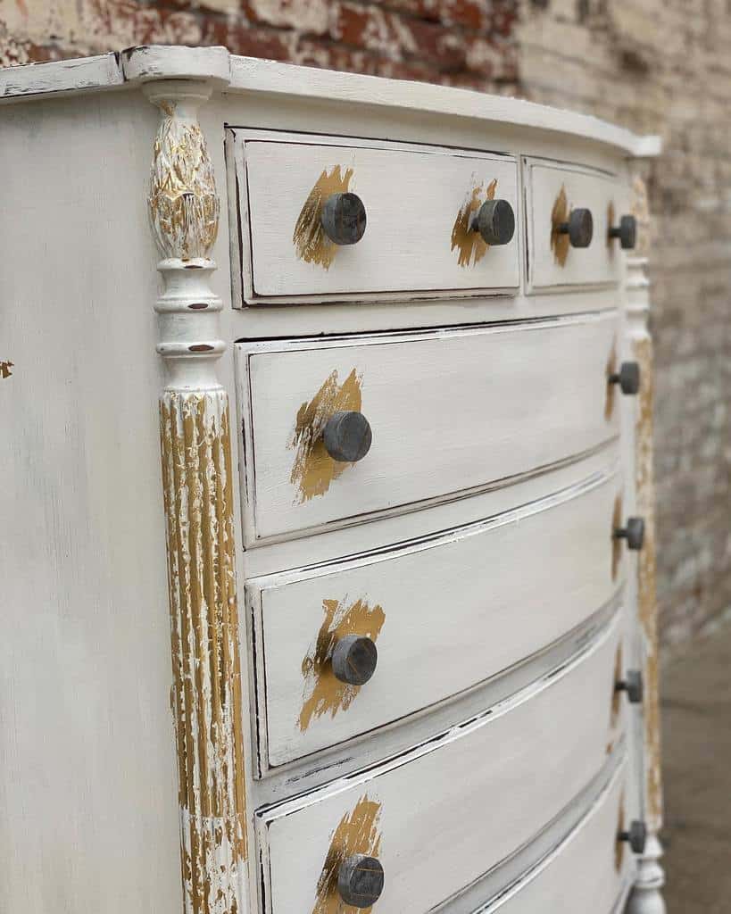 Distressed white chalk-painted dresser with exposed gold accents, dark round knobs, and ornate carved details against a rustic brick wall