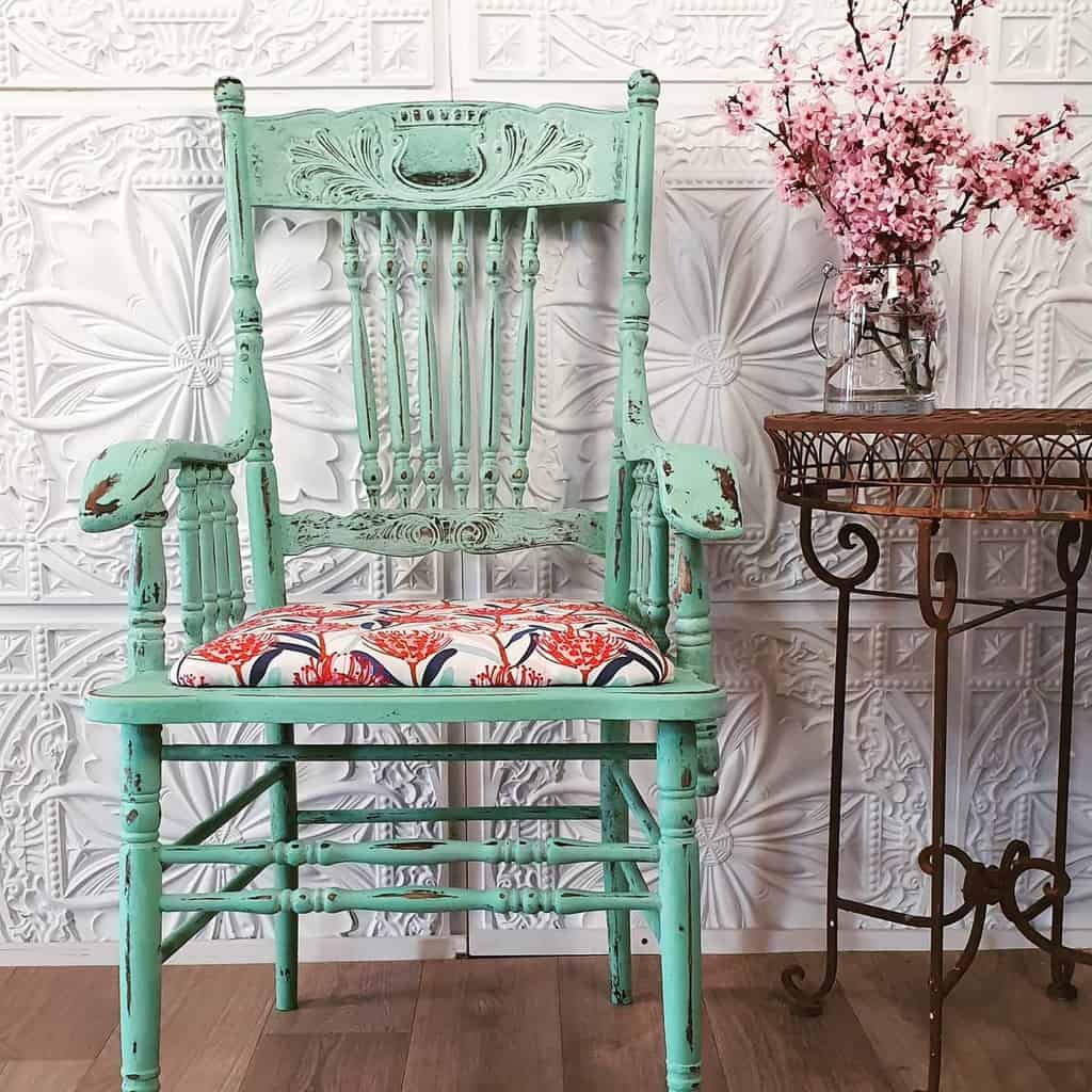 Distressed mint green vintage chair with intricate carvings and a floral cushion, styled with a rustic side table and pink blossoms